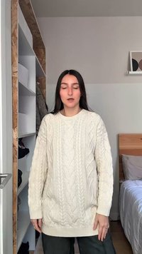 Femme aux yeux fermés portant un pull en maille torsadée blanc, debout dans une chambre avec un lit et des étagères en arrière-plan.