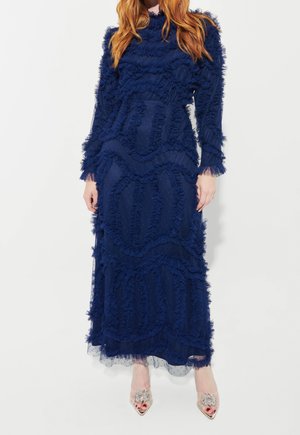 Femme portant une longue robe bleu marine texturée avec des détails à volants, debout, les mains détendues, chaussée de talons hauts transparents ornés de bijoux.