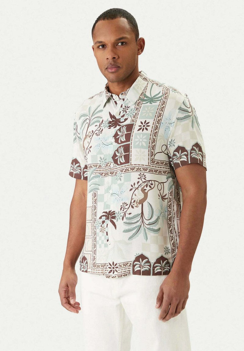 Homme portant une chemise à manches courtes avec des motifs floraux et géométriques verts et marron, associée à un pantalon blanc, sur un fond uni.