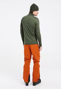 Grünes Langarmoberteil mit hohem Kragen, kombiniert mit orangen Skihosen mit seitlichen Reißverschlüssen und schwarzen Stiefelakzenten. Eine Mütze vervollständigt das Outfit.