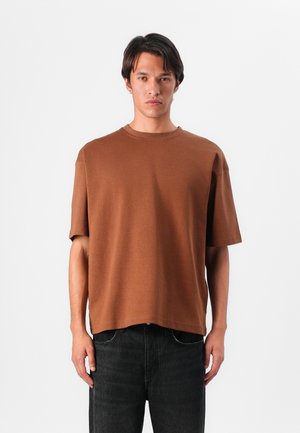 T-shirt oversize en coton brun avec des manches courtes, col rond et ourlet droit, porté avec un jean foncé. Texture lisse, design minimal.