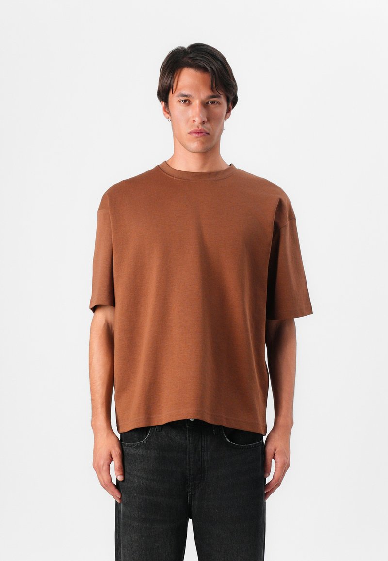 Hnedé bavlnené oversized tričko s krátkymi rukávmi, okrúhlym výstrihom a rovnými okrajmi, nosené s tmavými džínsami. Hladká textúra, minimalistický dizajn.