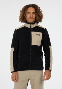 Hombre con cabello rizado que lleva una chaqueta de forro polar negra y beige con cremallera y pantalones beige, de pie contra un fondo blanco liso.