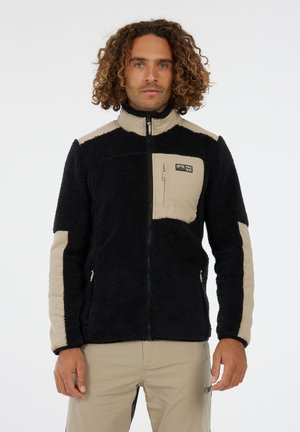 Man met krullend haar, gekleed in een zwart-beige ritsjack van fleece en beige broek, staande tegen een plain witte achtergrond.