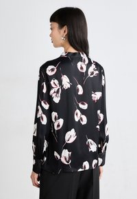 Svart bluse med blomstermønster i hvitt og rødt. Glatt stoff med lange ermer og belg med knapper. Skreddersydd design og avslappet passform.