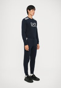 Tengerészeti pulóver és nadrág szett "EA" logóval; textúrált vállrész kiemelésekkel; karcsú fazon; fekete sportcipővel és fehér zoknival párosítva.