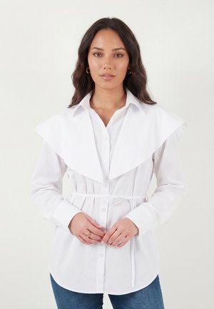 Naine laineliste tume juustega, kellel on seljas valge nööpidega särk, lai krae detaili ja vöös, seisab tavalise tausta ees.