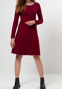 Vestido burdeos de manga larga con un cuerpo ajustado y falda acampanada. Hecho de una tela suave, cuenta con un escote redondo y un detalle sutil en la cintura.