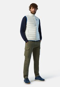 Gilet bianco piumoso con dettagli blu, camicia a maniche lunghe blu scuro, pantaloni cargo verdi e sneakers blu. Presenta cerniere e tasche laterali.