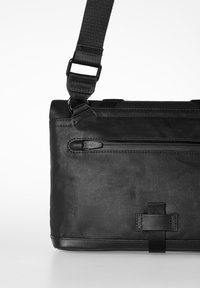 Black messenger bag with a textured fabric, adjustable strap, zippered front pocket, and a smooth leather accent at the bottom.