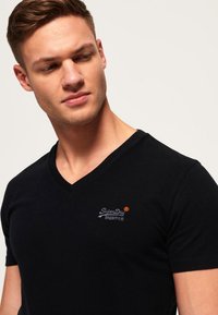 Camiseta de algodón negra con cuello en v que presenta un logo bordado en plata y naranja en el lado izquierdo del pecho. Textura suave y de mangas cortas.
