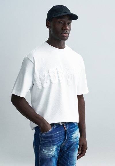 Λευκό βαμβακερό t-shirt με ανάγλυφη επιγραφή "ICON", σε συνδυασμό με ξεθωριασμένο μπλε τζιν. Το μοντέλο φοράει σκούρο καπέλο, με τα χέρια στις τσέπες.