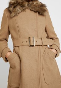Cappotto in lana cammello con colletto in pelliccia sintetica, vita con cintura e grandi tasche frontali. Presenta dettagli in metallo dorato e una finitura in tessuto testurizzato.