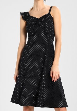 Zwarte mouwloze jurk met witte polkadots, aansluitend lijfje, gerimpelde band op één schouder en uitlopende rok boven de knie.