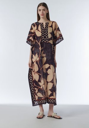 Young woman standing wearing a loose, ankle-length beige and dark purple patterned kaftan and beige sandals on a plain background.