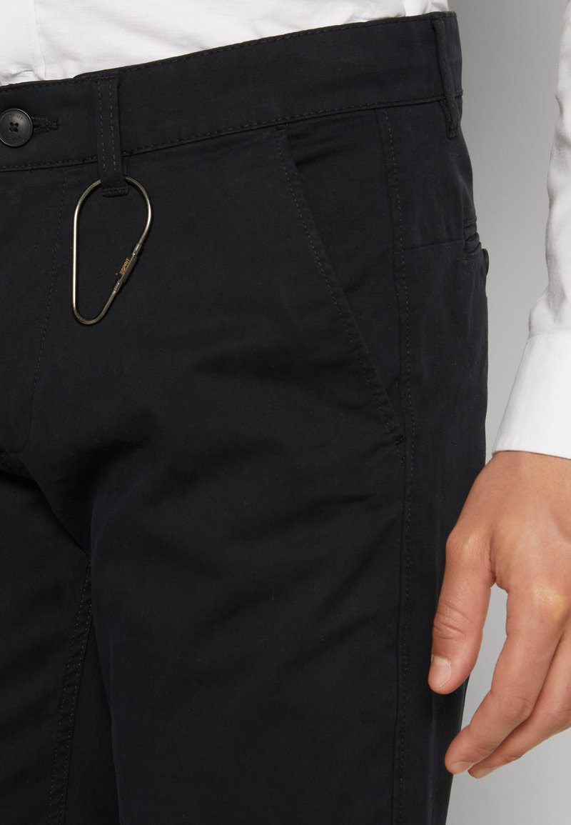 Pantalons noirs en coton avec une coupe slim, poches avant et un accent en mousqueton métallique sur la taille. Texture lisse et design épuré.