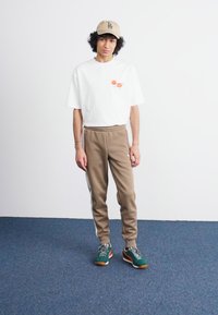 Tricou alb din bumbac cu un grafic mic portocaliu, pantaloni de trening bej, ușor tapered, și adidași verzi cu șireturi multicolore. Șapcă bej.