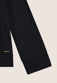 Top negro de manga larga con un acabado suave, que presenta detalles de ojetes bordados en los puños y una pequeña etiqueta de hardware dorado.