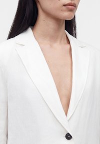 Κοντινό πλάνο ενός ατόμου που φοράει λευκό σακάκι με βαθύ V-λαιμόκοψη και ένα μαύρο κουμπί, δείχνοντας τον λαιμό, το στήθος και το κάτω μέρος του προσώπου.