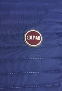 Marinblå pufferjacka i tyg med horisontella sömmar, med en rund maroon- och silverfärgad logotyp i form av ett märke med texten "COLMAR" på framsidan.