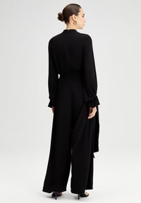 Salopetă neagră cu mâneci lungi, talie pliată și pantaloni largi. Material neted, opac, cu închidere prin fermoar la spate.
