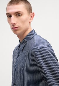 Μπλε σκούρο ριγέ πουκάμισο από λείο ύφασμα, με γιακά, κουμπί και μικρό κεντητό λογότυπο στο στήθος.