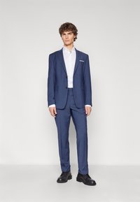 Marineblauer Anzug mit einem einreihigen Blazer und passender Hose, kombiniert mit einem weißen Hemd und schwarzen Schnürschuhen. Glatte Stoffoberfläche.