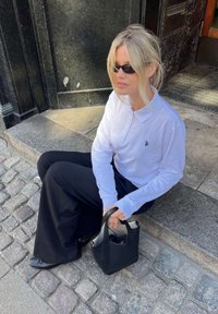 Blondīne, kas valkā saulesbrilles, baltu garroku polo kreklu un melnas platas bikses, sēž uz akmens kāpnēm, turot melnu rokassomu uz bruģa ielas.