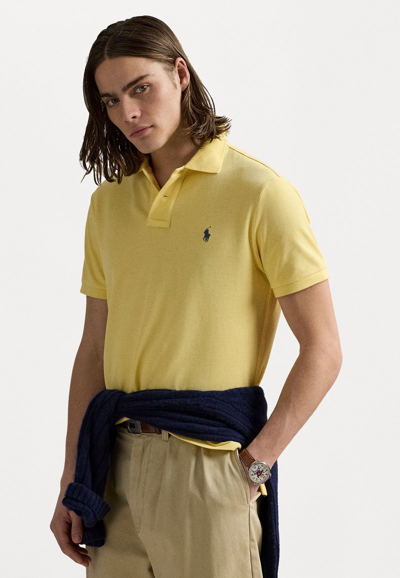 Cămașă polo galbenă, cu mâneci scurte, dintr-un material texturat, croială slim fit, cu un logo brodat bleumarin. Asortată cu pantaloni bej și un pulover bleumarin legat în talie.