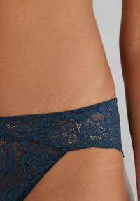 Marinblå spetsstring med intrikata blommönster, med scallopade kanter och en mjuk midjevidd som framhäver den delikata texturen.