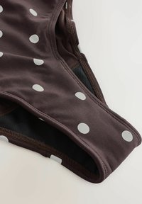 Nahaufnahme eines braunen Stoffes mit weißen Punkten und schwarzem Netzfutter, das die detaillierte Naht an einem Kleidungsstück zeigt.