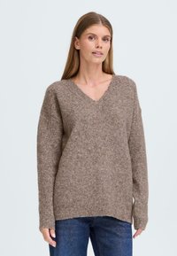 Brown knitted sweater with a V-neck, long sleeves, and ribbed hem. Soft texture and slightly oversized fit. Worn with blue jeans.