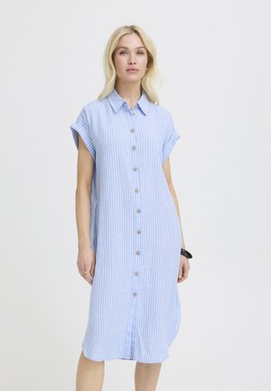 Heledate juustega naine, kes kannab põlvepikkust helesinist lühikeste varrukatega nööbitavat kleiti, millel on valged vertikaalsed triibud, seisab ühevärvilise tausta ees.