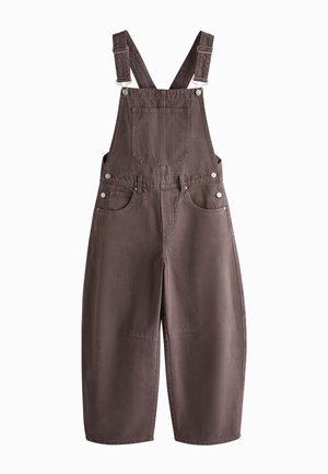 Persoon draagt een bruine denim tuinbroek over een wit T-shirt met korte mouwen met horizontale oranje en rode strepen, hand in de zak, gouden armband om de pols.