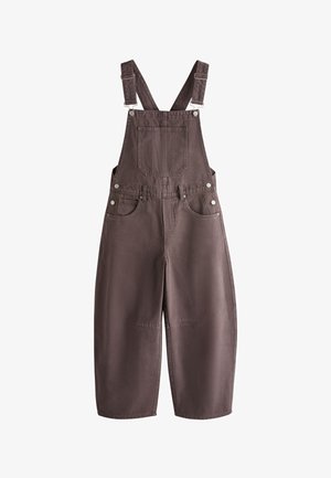 Persoană purtând un salopet maro din denim peste un tricou alb cu mânecă scurtă și dungi orizontale portocalii și roșii, mâna în buzunar, brățară de aur la încheietură.