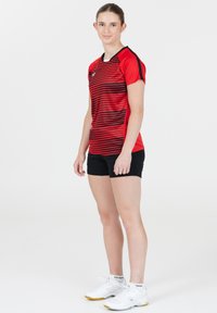 Rød og sort stribet atletisk skjorte med korte ærmer, parret med sorte shorts og hvide sneakers, på en model der står oprejst.