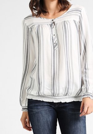 Vrouw draagt een witte blouse met lange mouwen met verticale grijze strepen, een geknoopte halslijn en een gerimpelde zoom, gecombineerd met een blauwe spijkerbroek.