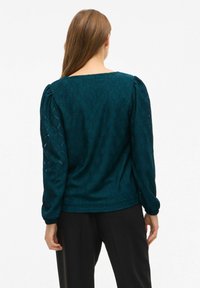Teal textuurblouse met een diamantpatroon, pofmouwen en een vierkante halslijn. Gecombineerd met zwarte broeken, die een relaxte pasvorm tonen.