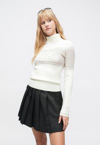 Hvit og beige strikkegenser med høyt halsanker, med teksten "Ski Club", kombinert med sort plissert skjørt. Myke, teksturerte materialer.