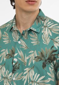 Camisa de manga corta en color teal con un estampado de hojas en tonos verdes y beige. Fabricada con tela texturizada, cuenta con botones blancos y un cuello.