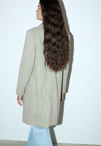 Bež plašč v houndstooth vzorcu z dolgimi rokavi, stranskimi žepi in ohlapnim krojem, oblečen čez svetlo modre široke hlače. Gladka tekstura.