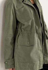 Chaqueta verde oliva hecha de un tejido duradero, con cierre de cremallera en el frente, cintura ajustable y dos bolsillos en el pecho con cierres a presión.