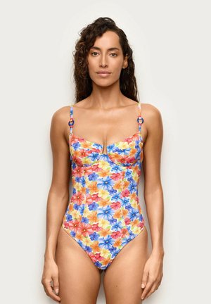 Mujer de pie vestida con un traje de baño de una pieza con un patrón floral colorido en azul, naranja, amarillo y rojo y tirantes delgados.