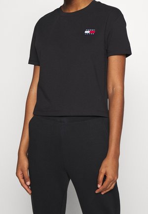 Persoon draagt een zwart kort mouw cropped t-shirt met Tommy Jeans-logo en bijpassende zwarte broek tegen een effen achtergrond.