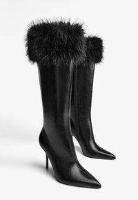 Čierne vysoké čižmy na ihlovom podpätku s ostrou špičkou, hladkým koženým materiálom a jemným čiernym kožušinovým lemom na vrchu. Obsahujú bočný zips.