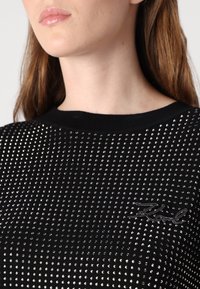 Juodas tekstūruotas megztinis su sidabrinėmis studelėmis, turintis raštą su briaunomis ir logotipo akcentą. Apskrito iškirpimo ir prigludęs stilius.