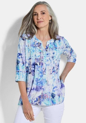 Femme mûre aux cheveux gris portant un chemisier à fleurs bleu et violet et un pantalon blanc, debout avec une main dans la poche.