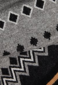 Écharpe tricotée présentant des motifs géométriques en gris et noir, comprenant des losanges et des zigzags, avec une finition douce et texturée.