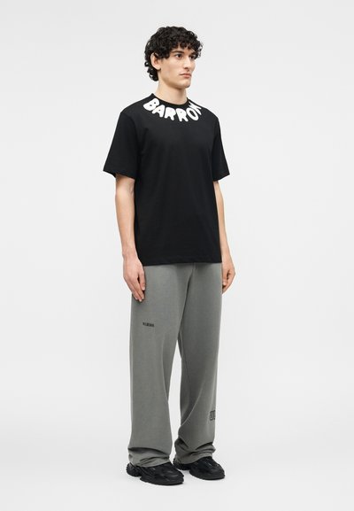 Νεαρός άνδρας όρθιος, φοράει μαύρο t-shirt με λευκό κείμενο γύρω από το γιακά και γκρι φαρδιά παντελόνια με μαύρα αθλητικά παπούτσια.