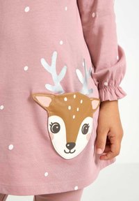 Różowa sukienka z długim rękawem w białe kropki, z kieszonką w kształcie brązowej twarzy jelenia zdobioną srebrnymi porożami i subtelnymi detalami twarzy.